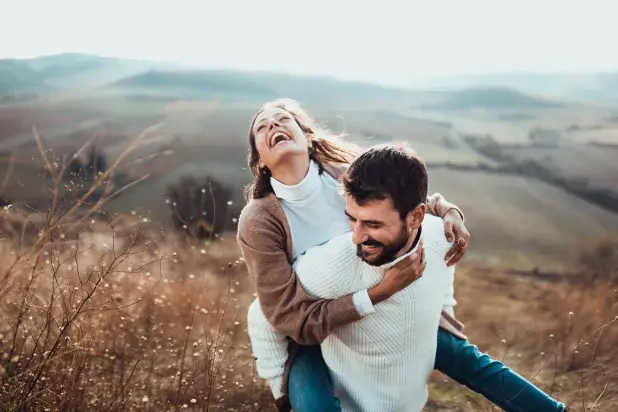 a smiling man giving a piggyback ride to a laughing woman on top of a hill with rolling countryside in the distance